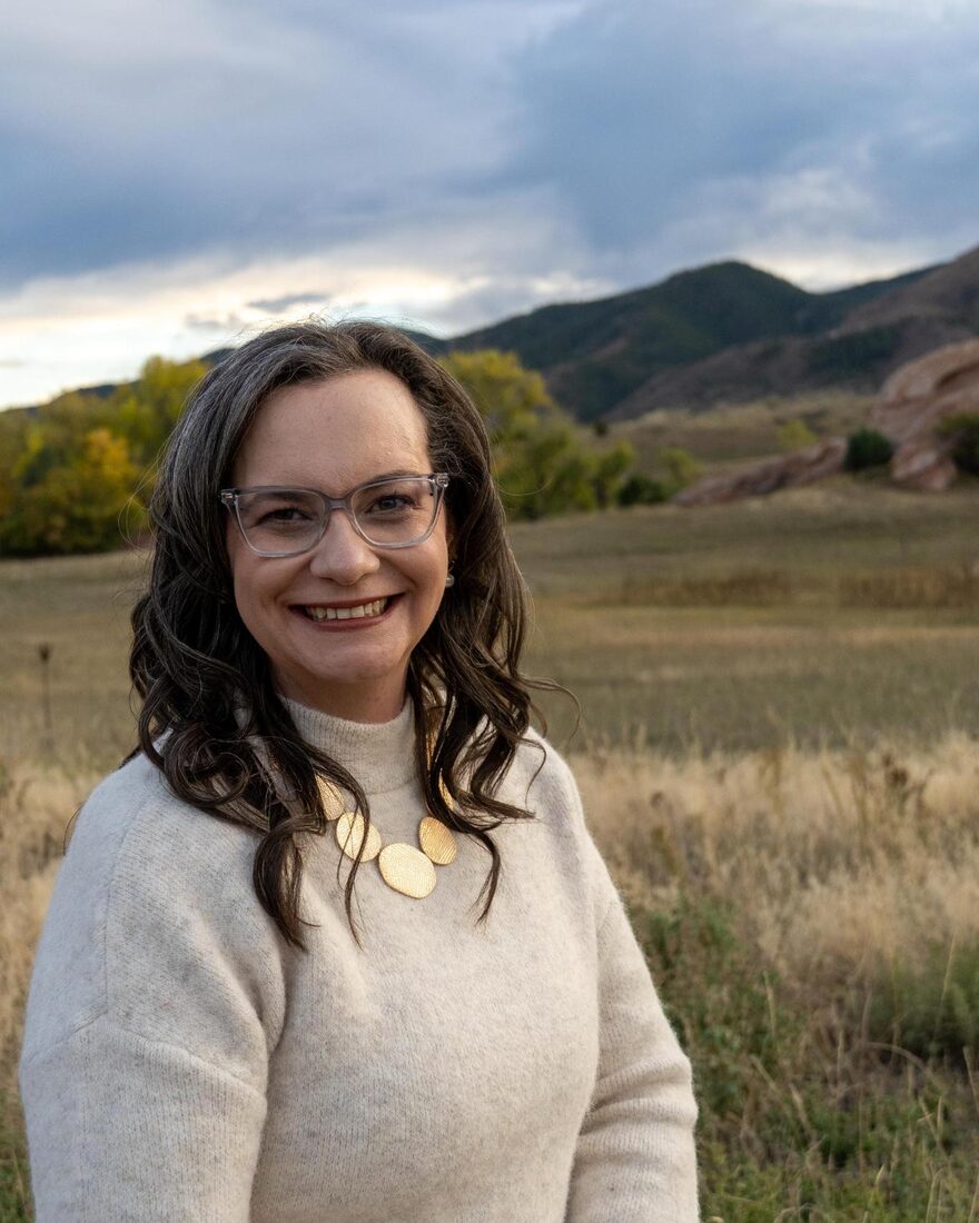 Sarah Otto, smiling, in a cream sweater and gold necklace, with a Colorado foothills meadow and red rock behind