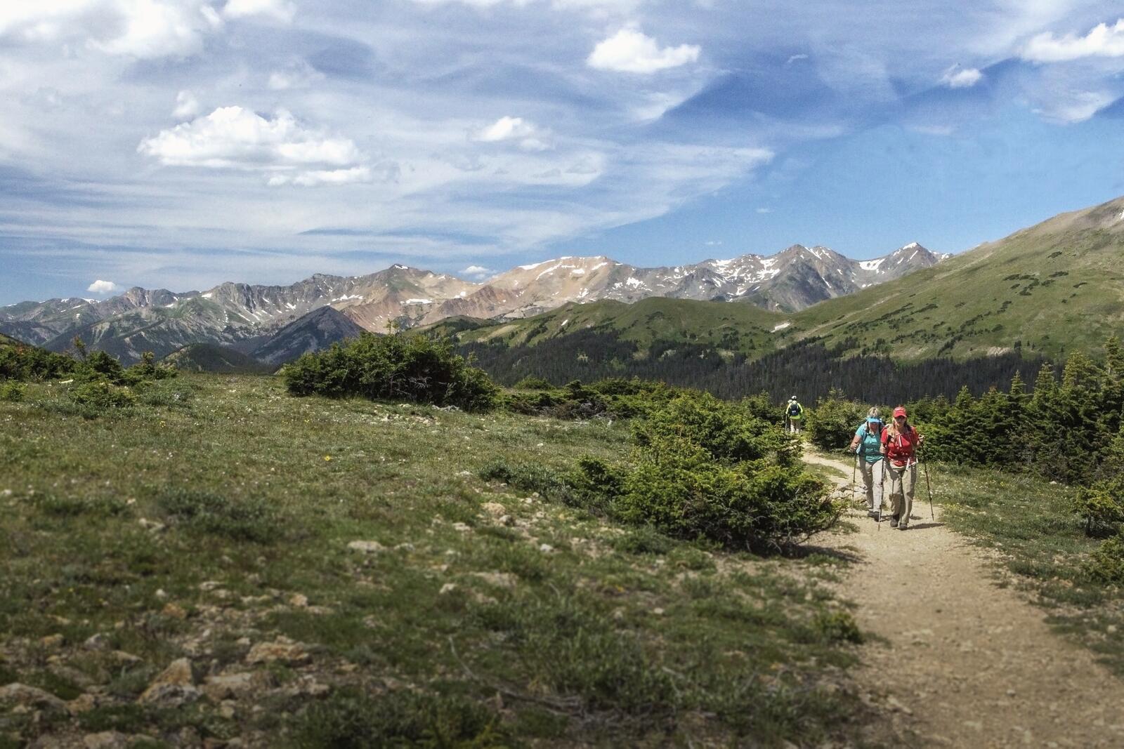 Two hikers with trekking poles climb a dirt trail across alpine tundra, snow-streaked peaks behind them